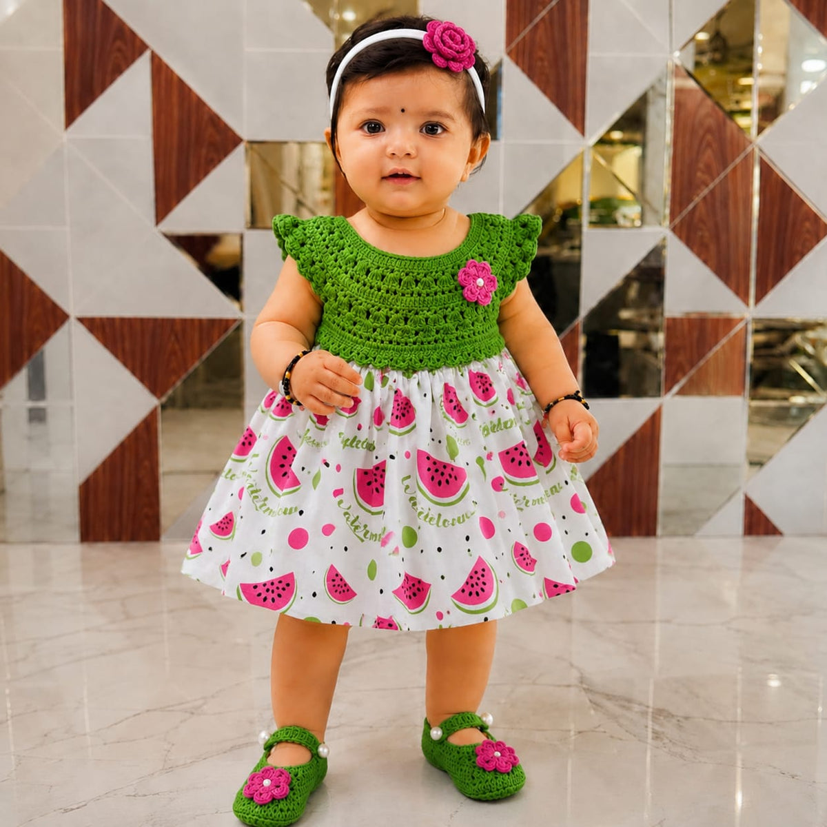 Baby wearing a green crochet-bodice frock with a white watermelon-print skirt, floral headband and matching shoes.