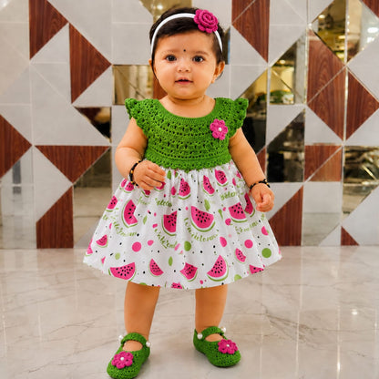 Baby wearing a green crochet-bodice frock with a white watermelon-print skirt, floral headband and matching shoes.